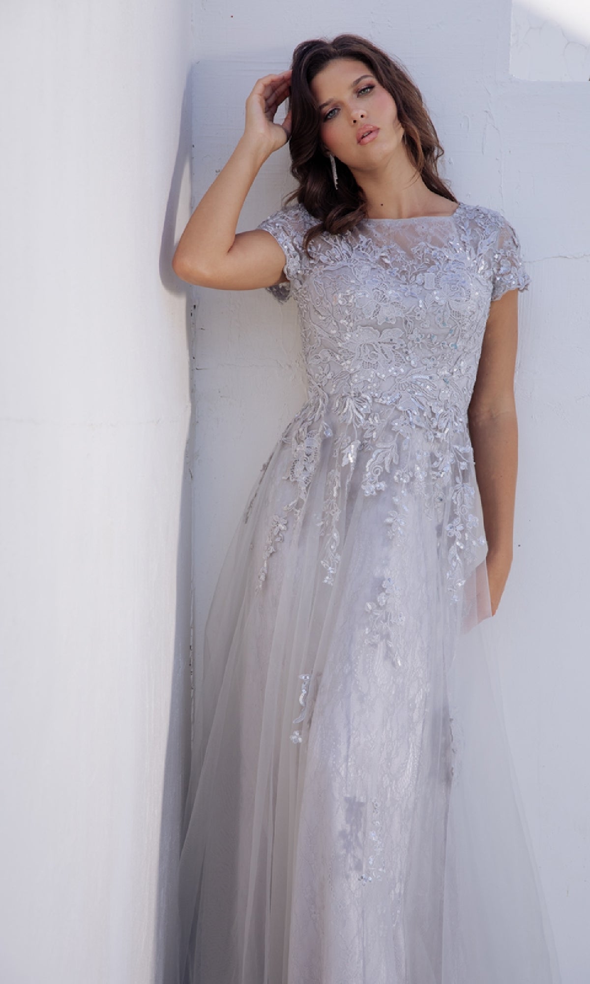 A woman wearing the Modest Short-Sleeve Prom Gown: Eureka EK104 poses against a white wall, facing the camera.